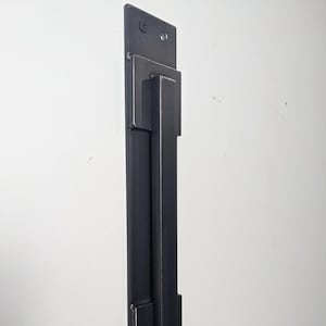 May include: Black metal door handle with a rectangular design. The handle is mounted on a vertical plate with two screws.