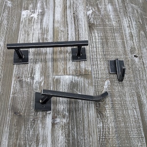 May include: A set of black metal bathroom fixtures, including a towel bar, toilet paper holder, and a hook. The fixtures are mounted on a weathered, gray-toned wooden surface, creating a rustic aesthetic.