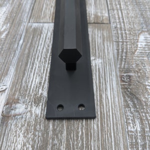 May include: Black metal door pull with a geometric, hexagonal handle. The pull is mounted on a rectangular plate with two pre-drilled holes for installation. The hardware has a matte finish and is designed for a modern aesthetic.