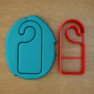 Peut inclure: Un emporte-pièce en plastique rouge en forme de porte-nom avec une empreinte bleu sarcelle de la même forme dans de l'argile à modeler.