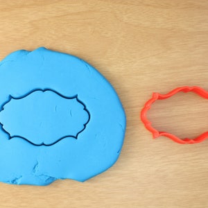 May include: A red plastic cookie cutter in the shape of a decorative frame, next to a blue piece of modelling clay with the same frame shape cut out.