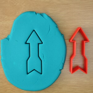 May include: A blue play dough with an arrow shape cut out of it. A red plastic arrow cookie cutter is next to the play dough.