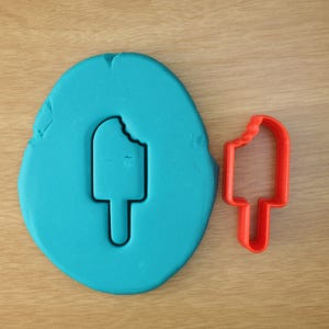 May include: A blue playdough shape with an ice pop impression and a red ice pop cookie cutter. The ice pop shape has a bite taken out of it. The objects are on a light brown wooden surface.