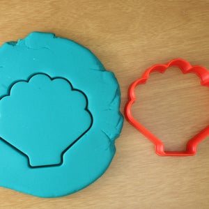 May include: A red plastic seashell cookie cutter on a teal blue background. The cookie cutter is shaped like a seashell with a scalloped edge.