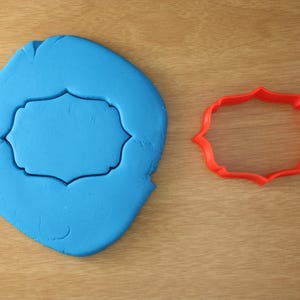 May include: A blue fondant cutout with a blank, rectangular space in the center. A red plastic cookie cutter with a similar shape is shown to the right.