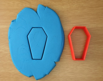May include: A blue fondant cutout of a coffin shape, next to a red plastic cookie cutter in the shape of a coffin.