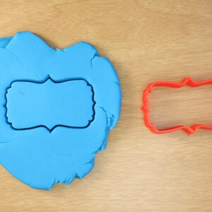 May include: A blue fondant heart with a red plastic cookie cutter in the shape of a decorative frame. The cookie cutter is sitting on a wooden surface.