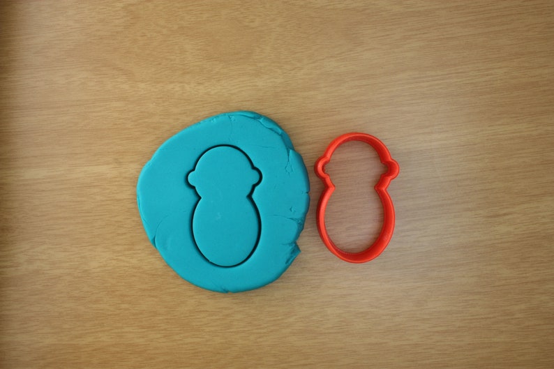 Puede incluir: Un cortador de galletas de pl&aacute;stico rojo con forma de mu&ntilde;eco de nieve est&aacute; junto a una bola de plastilina azul con una forma de mu&ntilde;eco de nieve presionada. Los art&iacute;culos est&aacute;n sobre una superficie de madera marr&oacute;n claro.