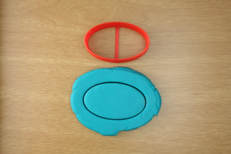 May include: A red plastic oval cookie cutter on a wooden surface, with a blue oval of modeling clay underneath it.