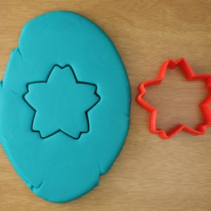 May include: A teal-colored playdough with a star-shaped cutout and a red star-shaped cookie cutter. The playdough is on a light brown wooden surface. The cookie cutter is made of red plastic.