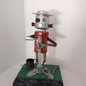 May include: A whimsical robot made from recycled materials, standing on a green base with a fork, a shovel, and a small bucket. The robot has a red torso, a silver head, and brown eyes.