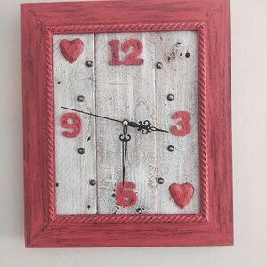Può includere: Un orologio con cornice rossa, legno dipinto di bianco e numeri dipinti di rosso. L'orologio ha due cuori rossi sul quadrante.