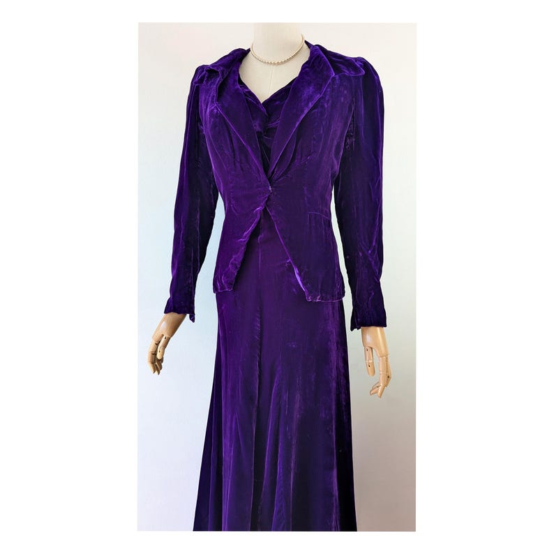 May include: A vintage, floor-length, purple velvet dress with a matching jacket. The jacket has a notched collar and long sleeves. The dress has a fitted bodice and a flowing skirt. The outfit is displayed on a mannequin. 1930s art deco