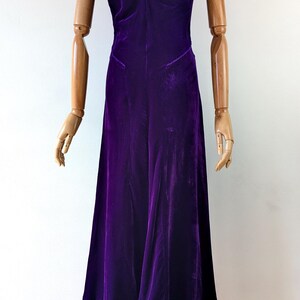 1930s art deco A full-length, deep purple velvet dress The garment has a draped neckline, fitted bodice long skirt. The dress is displayed on a wooden mannequin against a white background.
