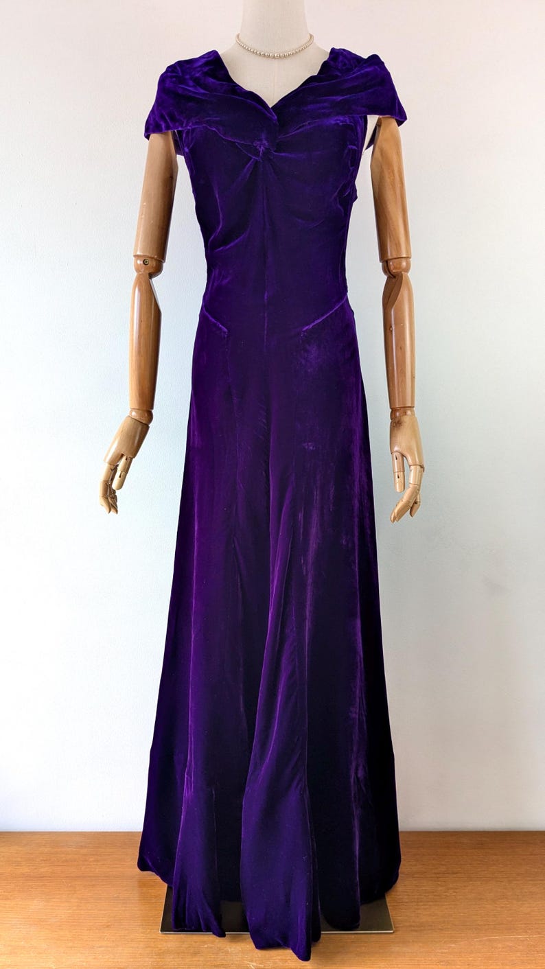 1930s art deco A full-length, deep purple velvet dress The garment has a draped neckline, fitted bodice long skirt. The dress is displayed on a wooden mannequin against a white background.