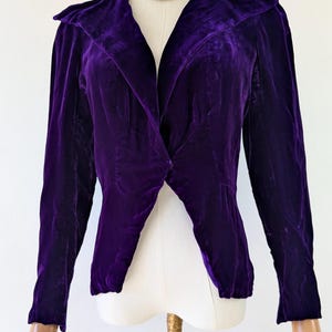 1930s art deco A vibrant purple velvet jacket with a shawl collar and long sleeves. The jacket is displayed on a mannequin, highlighting its tailored design and rich colour. The fabric appears soft and luxurious.