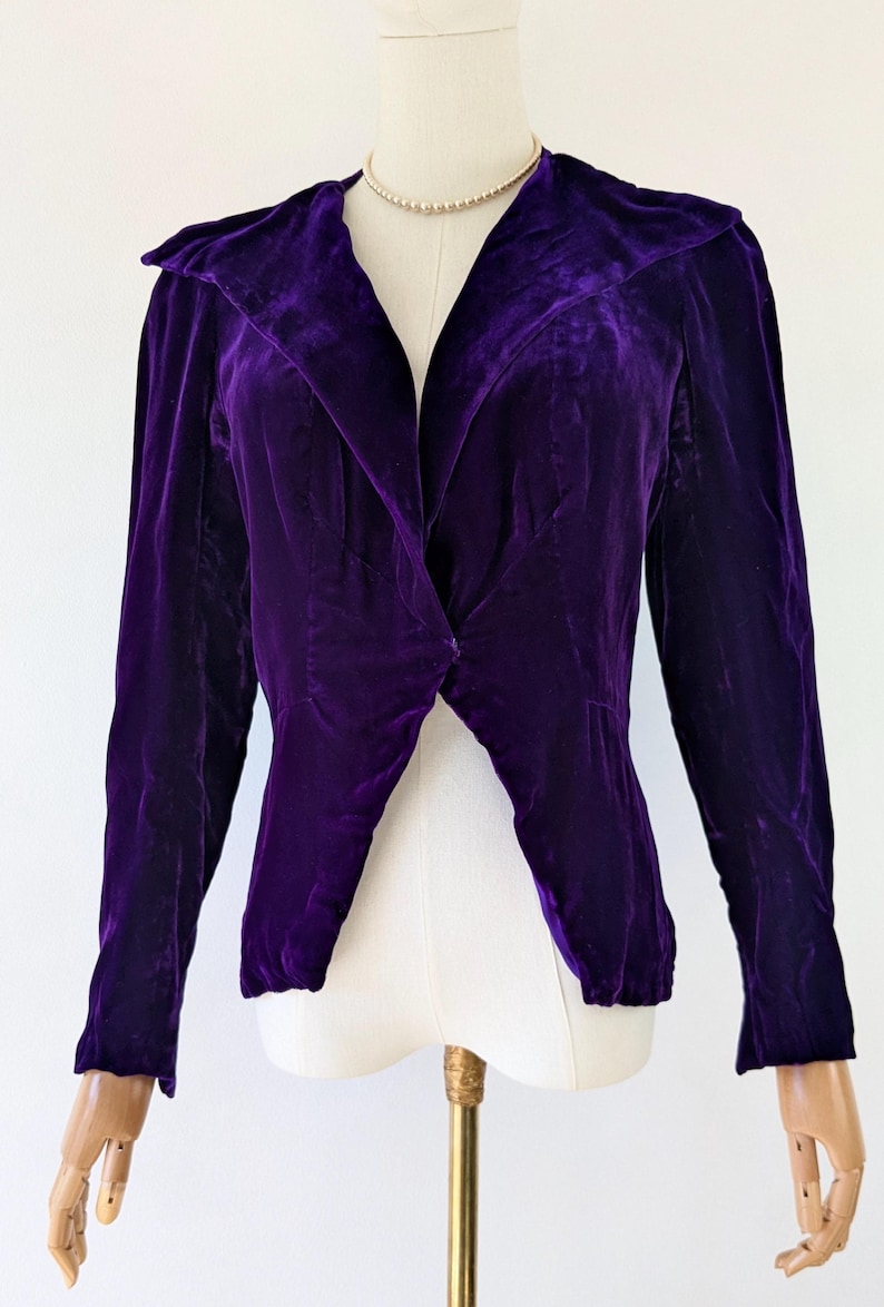 1930s art deco A vibrant purple velvet jacket with a shawl collar and long sleeves. The jacket is displayed on a mannequin, highlighting its tailored design and rich colour. The fabric appears soft and luxurious.