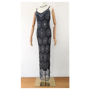 May include: A floor-length black sleeveless dress featuring a V-neckline and thin spaghetti straps. The garment is adorned with an intricate pattern of silver-toned circular beaded motifs and a beaded fringe detail along the neckline and hem.