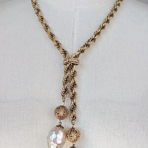 A vintage 1960s gold-tone lariat necklace featuring a thick, textured rope-style chain. The necklace is designed in a Y-shape, with the chain passing through a decorative slider set with a small, horizontal clear crystal. Two asymmetrical drops hang from the center, each featuring a large, shimmering baroque faux pearl suspended below an ornate, gold filigree ball bead. It is displayed on a white mannequin torso, highlighting its elegant, sophisticated "Mid-Century Glamour" aesthetic.