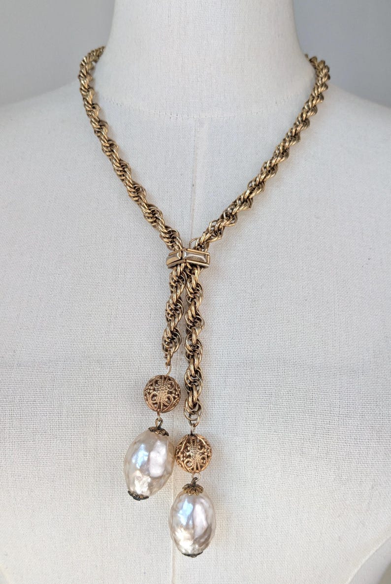 A vintage 1960s gold-tone lariat necklace featuring a thick, textured rope-style chain. The necklace is designed in a Y-shape, with the chain passing through a decorative slider set with a small, horizontal clear crystal. Two asymmetrical drops hang from the center, each featuring a large, shimmering baroque faux pearl suspended below an ornate, gold filigree ball bead. It is displayed on a white mannequin torso, highlighting its elegant, sophisticated "Mid-Century Glamour" aesthetic.