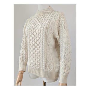 May include: Cream-colored cable-knit sweater with a mock neck. The sweater features various cable patterns across the front and sleeves. The garment is displayed on a mannequin, showcasing its texture and design. A classic, warm knitwear piece.
