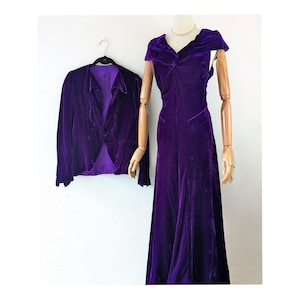1930s art deco A vintage purple velvet dress and matching jacket. The dress has a draped neckline and a flowing skirt. The jacket is a cropped style with a shawl collar. Both items are displayed on a mannequin against a white backdrop.