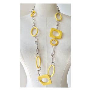 May include: A long necklace with a silver chain and yellow geometric shapes. The necklace features various shapes, including circles, ovals, and squares, all in a bright yellow colour. The necklace is displayed on a mannequin.