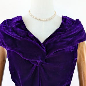 1930s art deco A close-up of a vintage, deep purple velvet dress. The dress features a draped neckline and short sleeves. A delicate pearl necklace adorns the mannequin's neck. The dress is displayed on a mannequin with wooden arms.