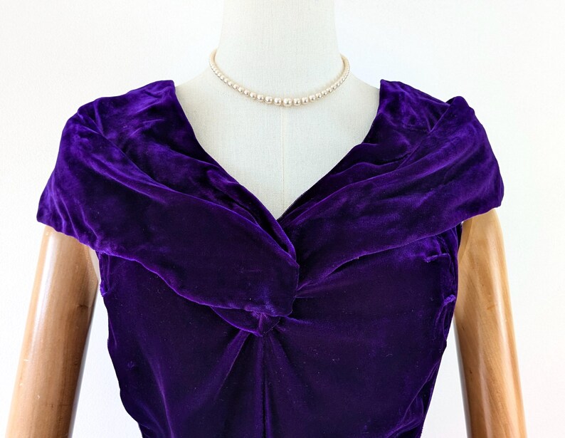 1930s art deco A close-up of a vintage, deep purple velvet dress. The dress features a draped neckline and short sleeves. A delicate pearl necklace adorns the mannequin's neck. The dress is displayed on a mannequin with wooden arms.