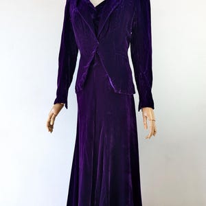 May include: A vintage, deep purple velvet suit displayed on a mannequin. The suit includes a long-sleeved blazer with a notched collar and a matching floor-length skirt. The fabric has a rich, lustrous sheen.