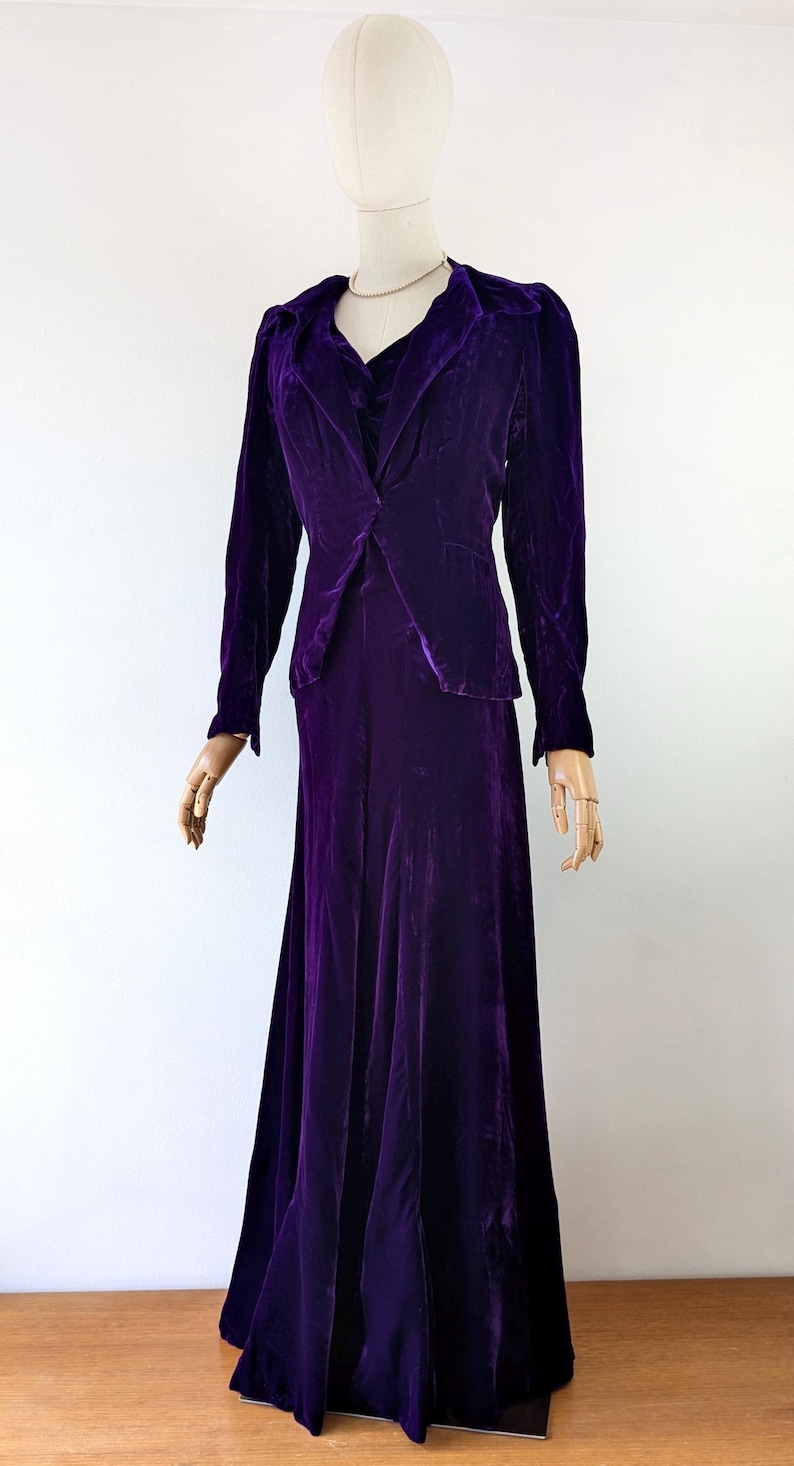 May include: A vintage, deep purple velvet suit displayed on a mannequin. The suit includes a long-sleeved blazer with a notched collar and a matching floor-length skirt. The fabric has a rich, lustrous sheen.