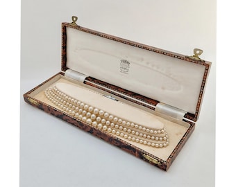 May include: A multi-strand pearl necklace rests inside an open, rectangular, faux tortoiseshell jewelry box. The interior lid features the text Lotus Pearls Made in England. The necklace is displayed on a cream-colored velvet insert.
