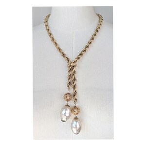 A vintage 1960s gold-tone lariat necklace featuring a thick, textured rope-style chain. The necklace is designed in a Y-shape, with the chain passing through a decorative slider set with a small, horizontal clear crystal. Two asymmetrical drops hang from the center, each featuring a large, shimmering baroque faux pearl suspended below an ornate, gold filigree ball bead. It is displayed on a white mannequin torso, highlighting its elegant, sophisticated "Mid-Century Glamour" aesthetic.