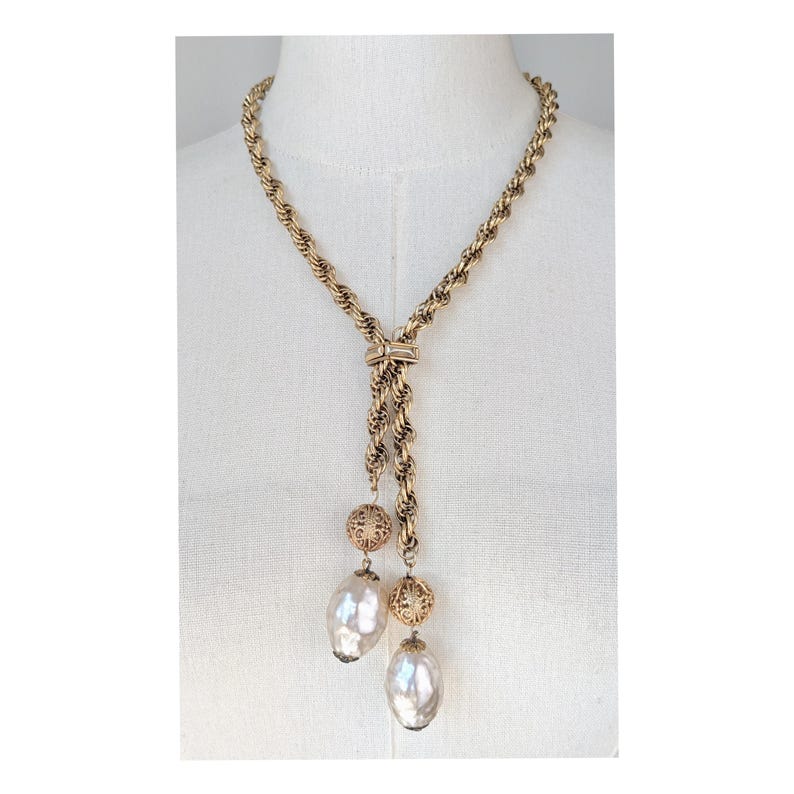 A vintage 1960s gold-tone lariat necklace featuring a thick, textured rope-style chain. The necklace is designed in a Y-shape, with the chain passing through a decorative slider set with a small, horizontal clear crystal. Two asymmetrical drops hang from the center, each featuring a large, shimmering baroque faux pearl suspended below an ornate, gold filigree ball bead. It is displayed on a white mannequin torso, highlighting its elegant, sophisticated "Mid-Century Glamour" aesthetic.