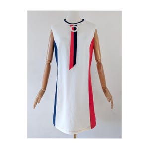 May include: A sleeveless white shift dress featuring a round neckline with a dark blue trim. The dress includes a vertical red and dark blue necktie detail secured by a white circular ring at the collar, with solid dark blue and red side panels.