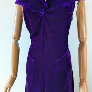May include: A deep purple velvet dress with a draped neckline and fitted bodice. The dress is displayed on a wooden mannequin, showcasing its elegant design and flowing silhouette. The dress appears to be vintage or retro.