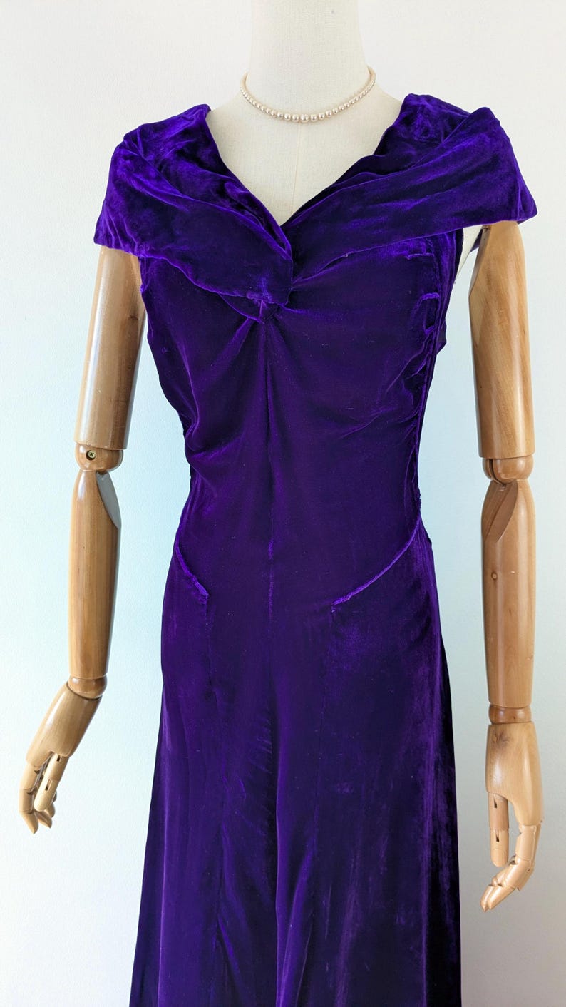 May include: A deep purple velvet dress with a draped neckline and fitted bodice. The dress is displayed on a wooden mannequin, showcasing its elegant design and flowing silhouette. The dress appears to be vintage or retro.