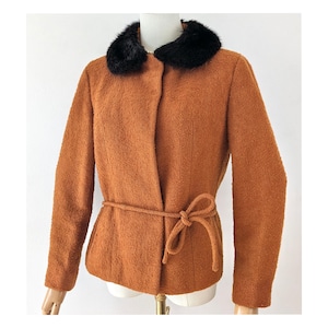 May include: A textured burnt orange jacket featuring a contrasting black faux fur collar and a matching fabric tie belt at the waist. The jacket has long sleeves and a clean, buttonless front closure, providing a structured yet soft appearance.