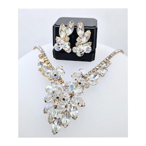 Könnte beinhalten: Ein goldfarbenes Halsketten- und Ohrring-Set. Der Schmuck besteht aus klaren, facettierten Kristallen, die in floralen und blattartigen Mustern angeordnet sind. Die Ohrringe werden auf einer schwarzen Box präsentiert. Die Halskette hat eine zentrale Kristallansammlung.