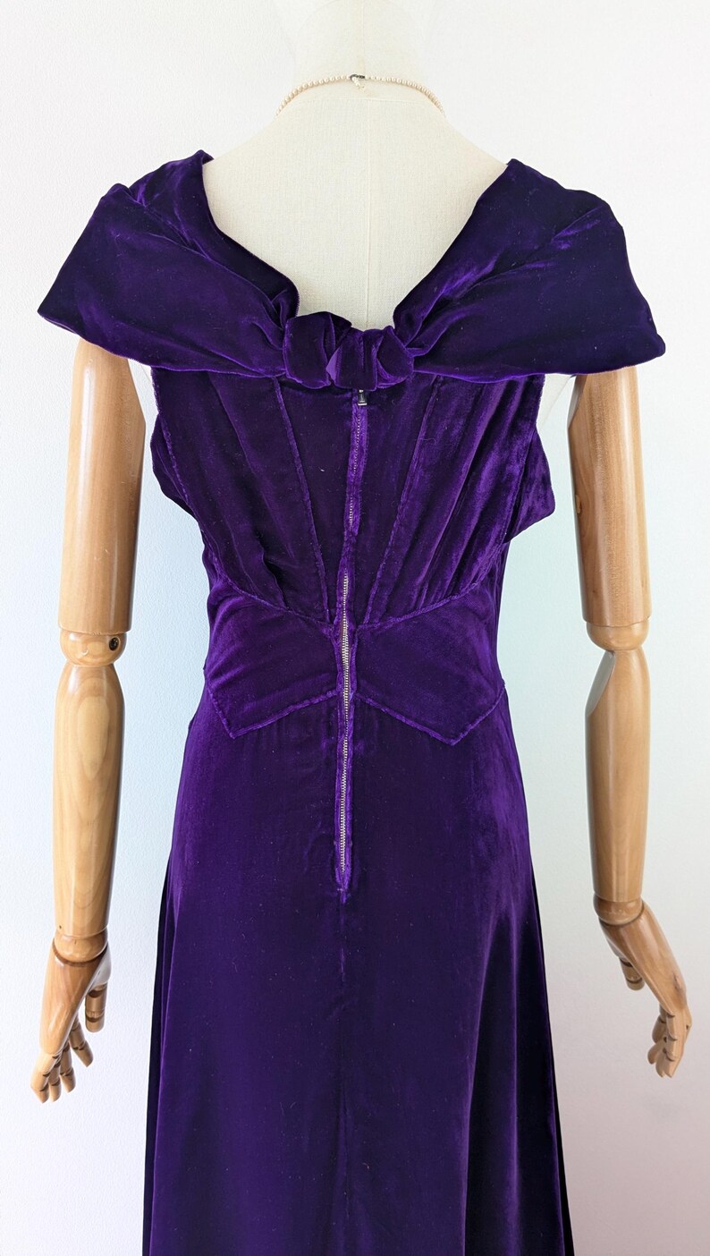 May include: A vintage, sleeveless, purple velvet dress. The dress features a bow detail at the back and a full-length zipper. The dress is displayed on a mannequin.
