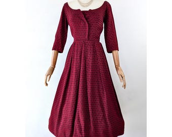 May include: A vintage burgundy dress with a textured pattern. The dress features a boat neckline, three-quarter sleeves, and a fitted waist that flows into a flared skirt. The dress is displayed on a mannequin with a pearl necklace.