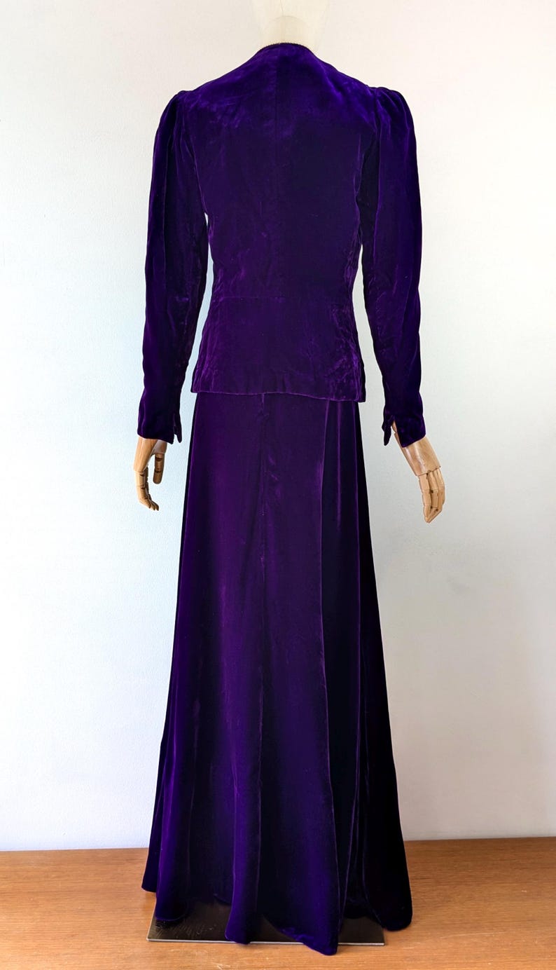 May include: A vintage, two-piece, purple velvet suit. The jacket has long sleeves and a tailored fit, paired with a matching floor-length skirt. The rich color and luxurious fabric suggest a formal or evening wear ensemble.