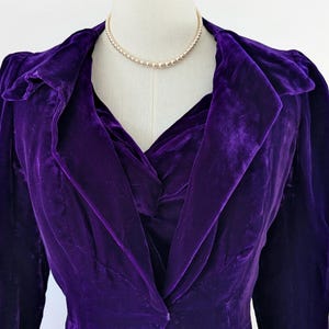 May include: A close-up of a vintage purple velvet jacket on a mannequin. The jacket has a shawl collar and long sleeves. A pearl necklace is draped around the mannequin's neck. The jacket's fabric has a rich, textured appearance.