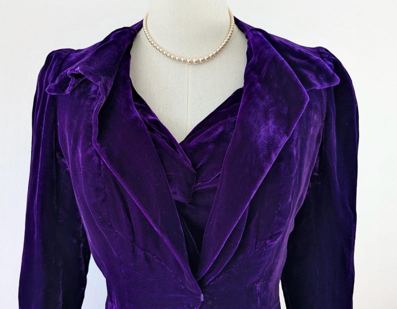 May include: A close-up of a vintage purple velvet jacket on a mannequin. The jacket has a shawl collar and long sleeves. A pearl necklace is draped around the mannequin's neck. The jacket's fabric has a rich, textured appearance.