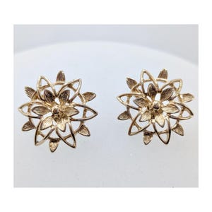 Może przedstawiać: Para złotych kolczyków sztyftów. Każdy kolczyk ma kwiatowy wzór z wieloma płatkami i centralnym kwiatem. Kolczyki są umieszczone na białym tle.
