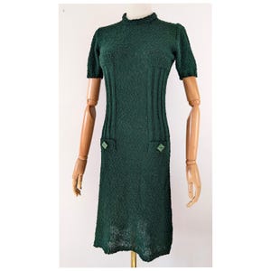 May include: A dark green knee-length dress with a textured, crinkled fabric surface. The garment features a high mock neckline, short sleeves, and two vertical ribbed panels on the front bodice. Two small, square, light green buttons accent the front.