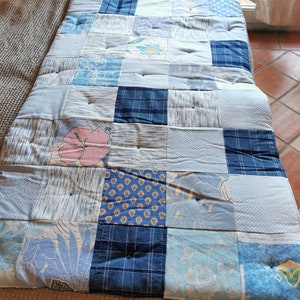 Peut inclure: Une courtepointe en patchwork réalisée à partir de différents tissus bleus et blancs. La courtepointe est composée de carrés de motifs différents, notamment floraux, à carreaux et géométriques. La courtepointe est matelassée avec un point simple et a un dos bleu clair.