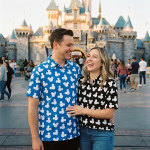 May include: A couple smiles in front of a castle. The man wears a blue button-down shirt with white Mickey Mouse patterns. The woman wears a black shirt with white Mickey Mouse patterns and blue jeans. The background features a castle and other people.