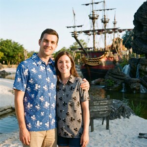 Könnte beinhalten: Ein Paar posiert vor einem Piratenschiff. Der Mann trägt ein blaues Hemd mit weißem Mickey-Mouse-Muster, kombiniert mit einer khakifarbenen Hose. Die Frau trägt ein graues Hemd mit schwarzem Mickey-Mouse-Muster.