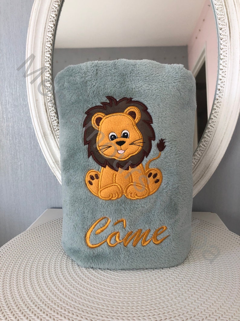 May include: A soft gray plush blanket with an embroidered lion design and the name "Come" in gold thread.